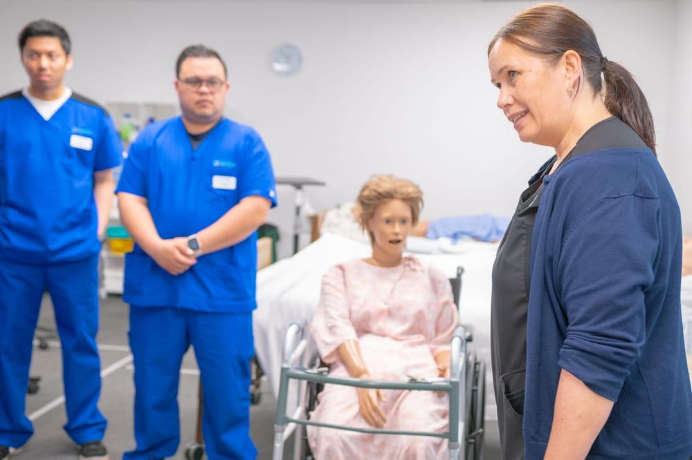 An instructor in a simulation lab talking to two students in scrubs beside a patient mannequin seated in a wheelchair