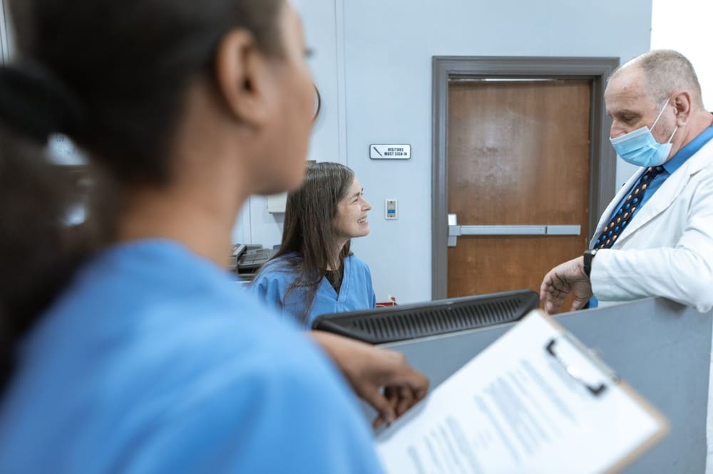 Two health professionals in scrubs and a lab coat conferring over a folder at a hospital reception desk