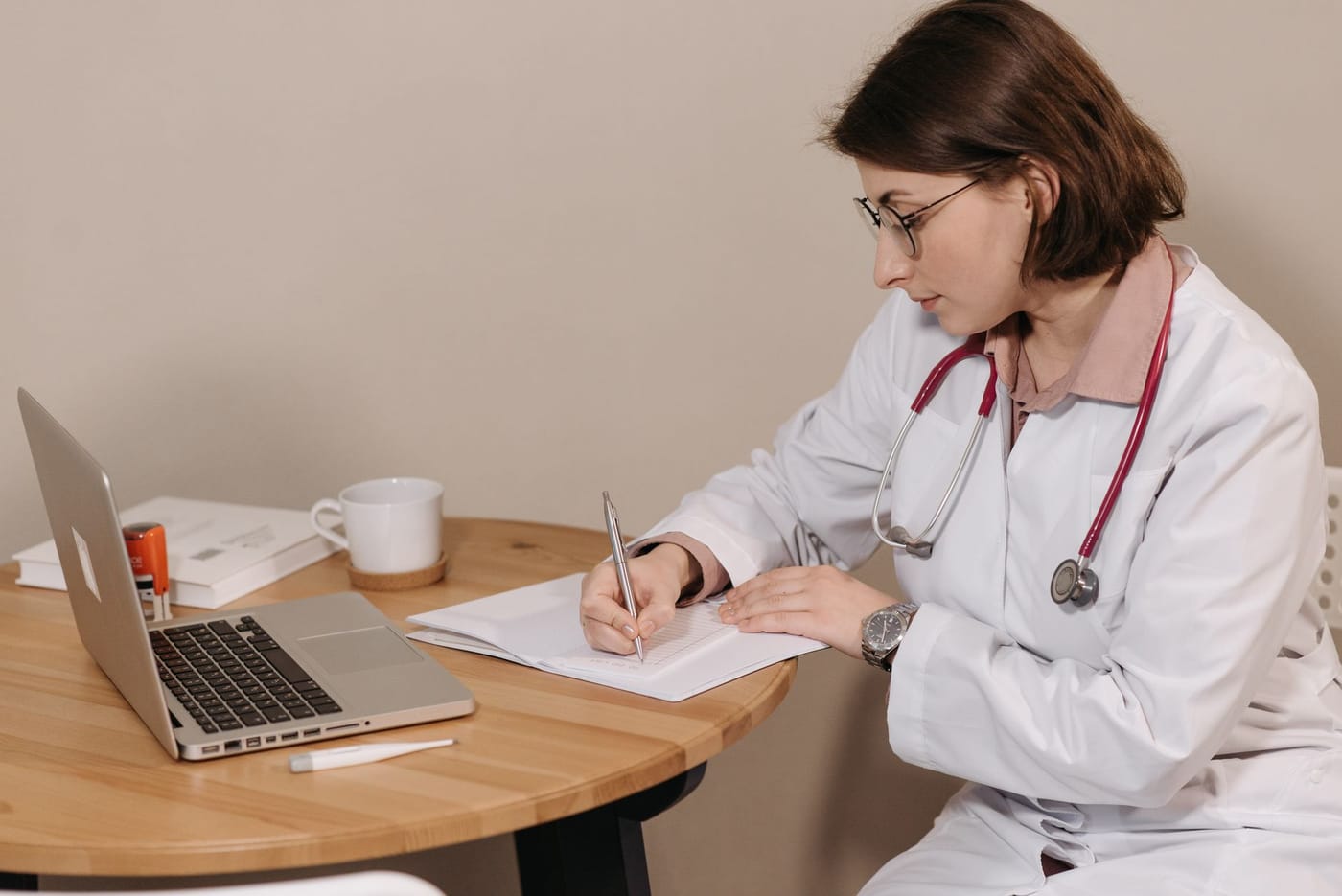 A clinician in a white coat and stethoscope writing notes beside an open laptop and a stack of textbooks